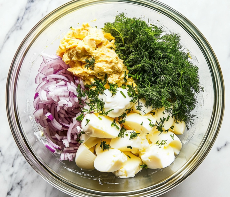 A glass mixing bowl containing chopped red onions, boiled eggs, fresh dill, and a creamy yellow mixture on a marble countertop.