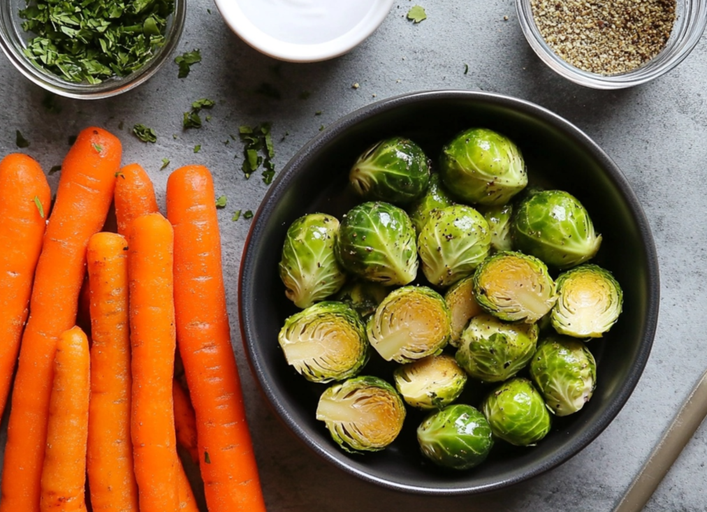 Fresh carrots and seasoned Brussels sprouts in a black bowl, surrounded by herbs, salt, and pepper.